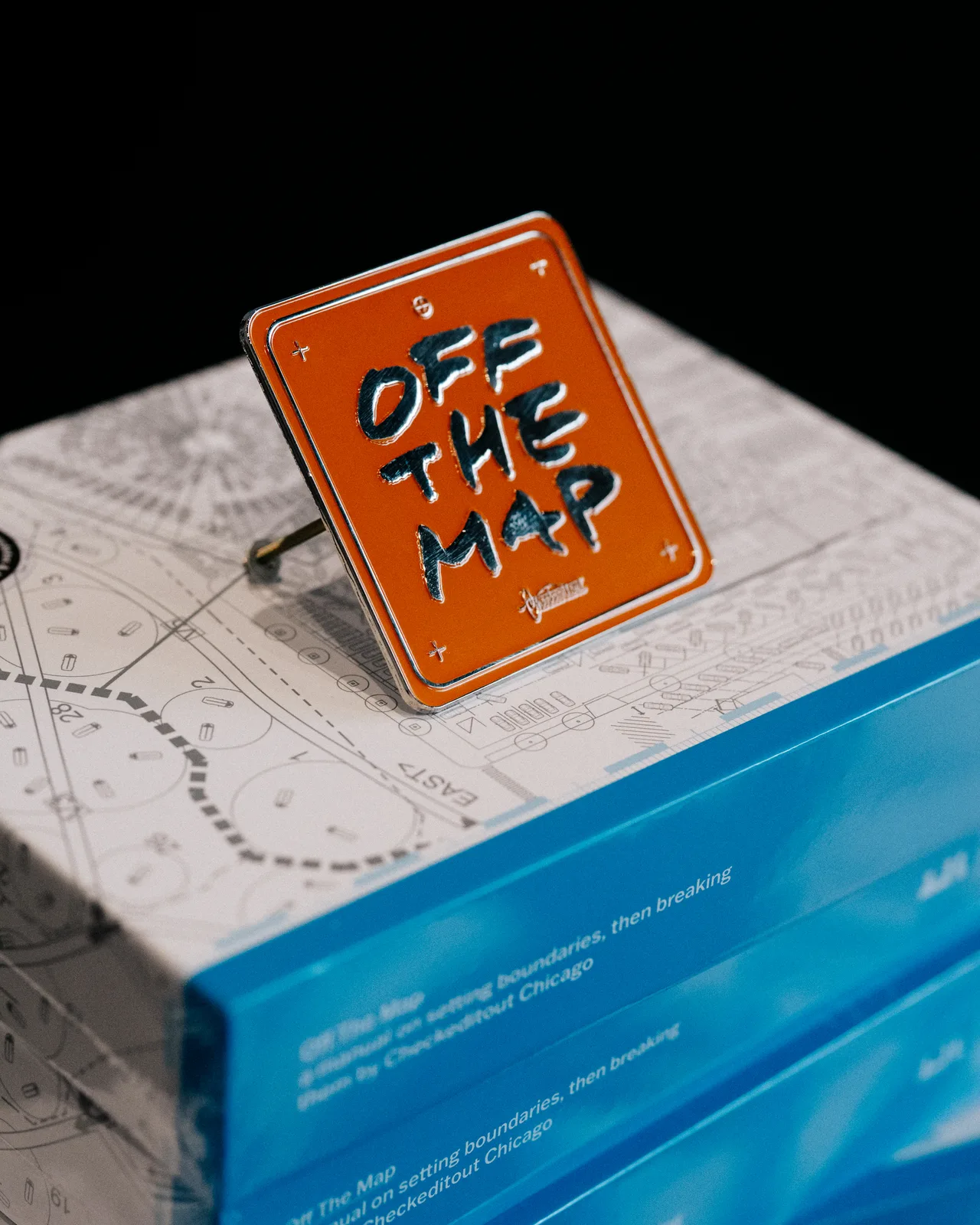 An orange Off the Map pin resting on the book and its route illustration
